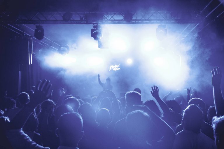 A crowded concert with people raising their hands, silhouetted against blue stage lights and smoke under a lit sign.