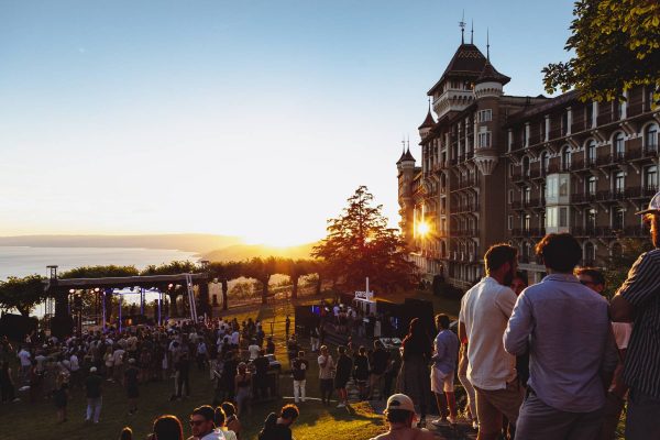 Outdoor event at sunset with people gathering on a lawn near a stage with lighting equipment, overlooking a lake, beside a grand historic building with multiple turrets.