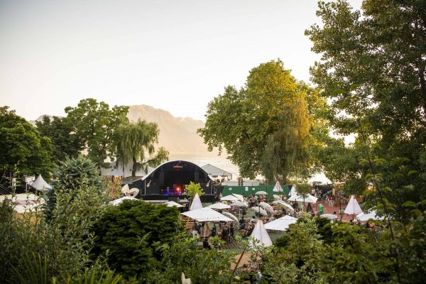 Outdoor festival scene with a stage, audience under sunshades, surrounded by greenery, with mountains and a body of water in the background.