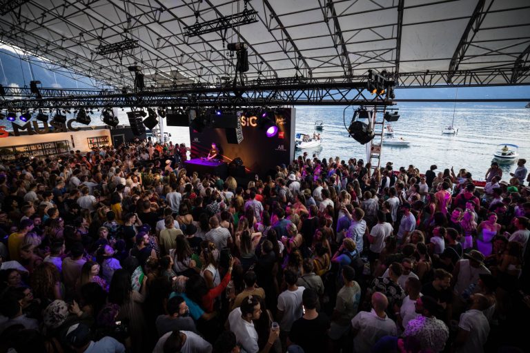 A large crowd of people gathered at an outdoor music event with a DJ performing on stage, set against a backdrop of the ocean with boats visible in the distance.