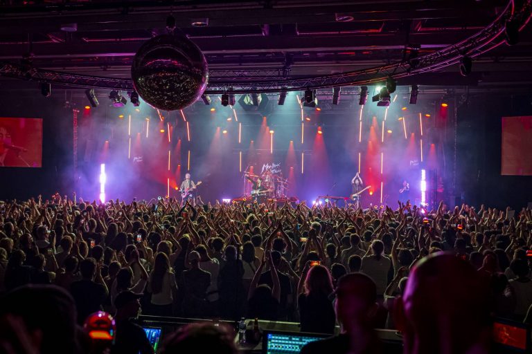 A live concert scene with The Rose performing on stage, vibrant stage lights casting colorful beams, a large disco ball hanging overhead, and a crowd of fans raising their hands in excitement.