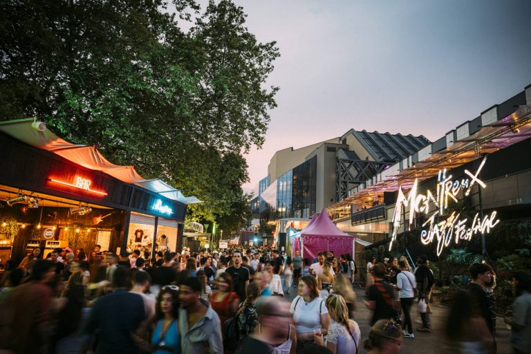 A bustling outdoor evening street scene with a crowd of people, some blurred in motion, between stalls and a neon sign for a jazz festival.