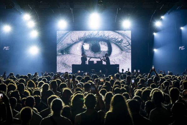 A concert scene with a crowd facing a stage where The Blaze perform, backlit by spotlights and in front of a large screen displaying a close-up of an eye.