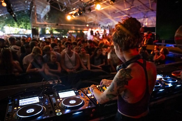 Noria Lilt with tattoos is operating a mixing console at a vibrant night event, with an attentive crowd in the background under illuminated tents.