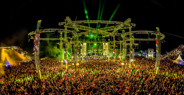 An expansive outdoor music festival at night with a large crowd of people, a stage with bright lights and lasers illuminating the scene.