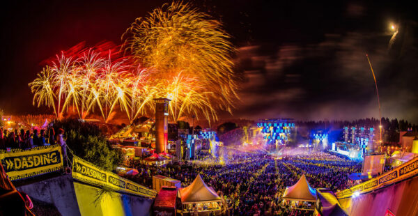 A panoramic view of a vibrant outdoor music festival at night with a massive crowd, illuminated stages, surrounding tents with logos, and a spectacular fireworks display in the sky.