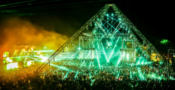 A crowded outdoor music festival at night with a large, illuminated triangular stage structure and dynamic green stage lights.