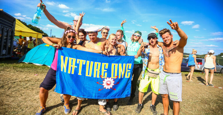 A group of lively young adults holding up a blue 'NATURE ONE' flag at a festival, with tents in the background and a clear blue sky above. Some are making peace signs and wearing casual summer clothing.