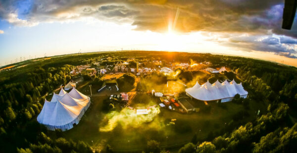 Aerial view of an outdoor festival with multiple tents and structures, surrounded by trees at sunset with rays of sunlight shining through a partly cloudy sky.