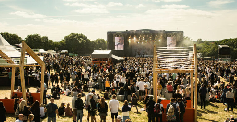 Outdoor music festival scene with a large crowd of people in front of a stage, some seated on the grass, others standing, with clear skies overhead and festival structures scattered around.