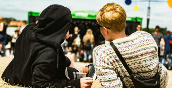 Two people sitting and chatting at an outdoor festival with others in the background and a stage visible in the distance. One person is wearing a black hoodie, and the other has a patterned sweater.
