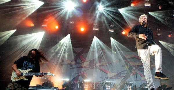 A live concert performance with dynamic lighting; a guitarist on the left with long hair covering his face playing an electric guitar, and a lead singer on the right with dyed hair, gesturing mid-performance, with a backdrop displaying abstract graphics. Lights beam down from above the stage.