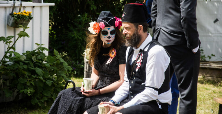 A woman in a polka dot dress wearing a Day of the Dead mask and a man in a traditional black vest and hat seated outside, both holding paper cups, with plants and a white structure in the background.