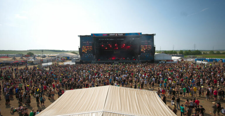 An outdoor music festival with a large crowd in front of a stage with 'Nova Rock' banners, in a sunny, open field setting.