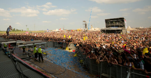 A large crowd gathered at an outdoor music festival with people standing near a stage, some holding up flags, and security personnel in the foreground.