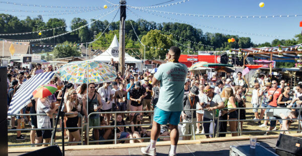 A vibrant outdoor festival scene with a diverse crowd of people, some holding drinks and umbrellas for shade, enjoying a performance on a sunny day with string lights above. A man with his back to the camera stands on the stage, addressing the audience.