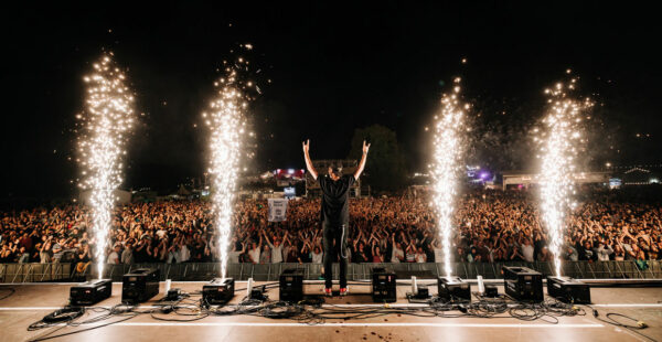 A performer raises their arms triumphantly in front of a large, cheering crowd at an outdoor concert, with bright pyrotechnics flaring on either side of the stage.