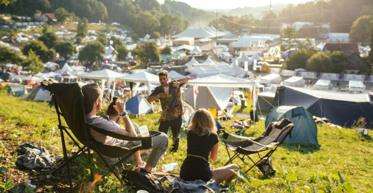 Group of people relaxing in camping chairs on a grassy hill overlooking a crowded festival campground with tents and structures in the background during sunset.