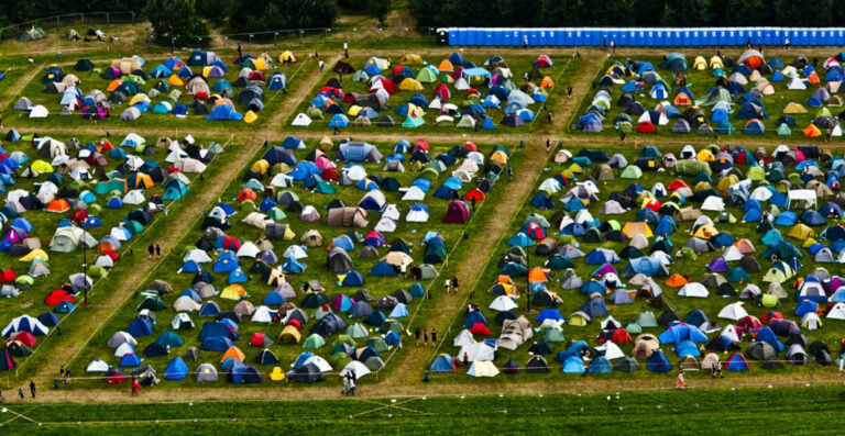 Aerial view of a crowded festival camping area with rows of colorful tents neatly arranged on a grassy field, with walkways in between and portable toilets lined up along the top edge.