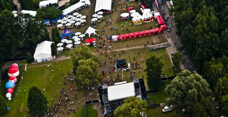Aerial view of an outdoor festival with tents, food stalls, and a crowd of people surrounded by trees.