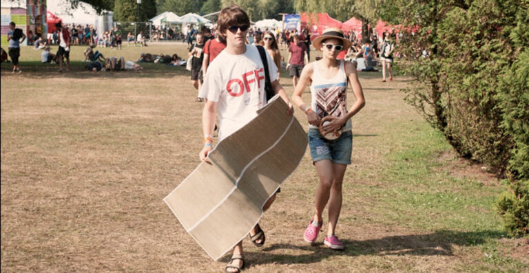 Two people walking across a grassy area at a festival, one carrying a folded camp bed and the other holding a ball, with festival-goers in the background.