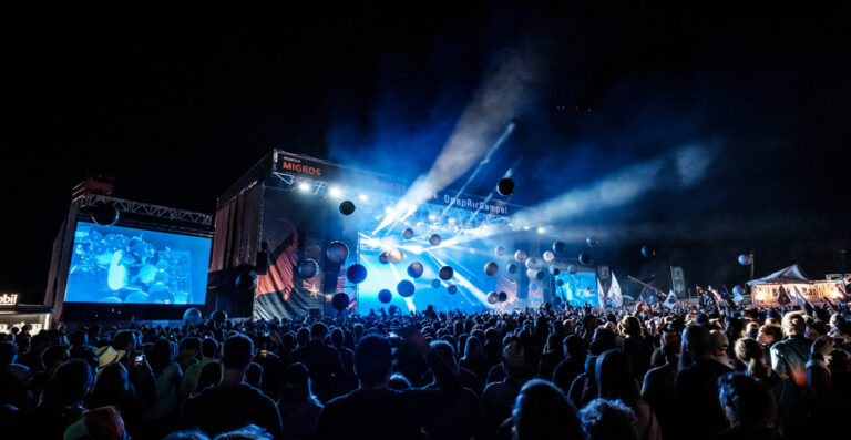 Crowd of people at an outdoor music festival at night with stage lights, a large screen, and flying balloons.