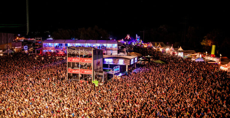 Aerial nighttime view of a densely packed outdoor music festival crowd with lit-up stages and festival booths.