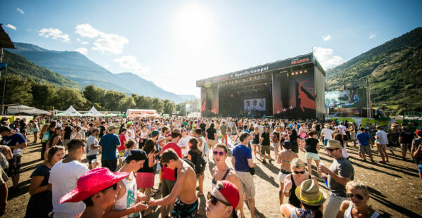 Crowd of people enjoying an outdoor music festival with a stage in the background set against a backdrop of mountains under a sunny sky.