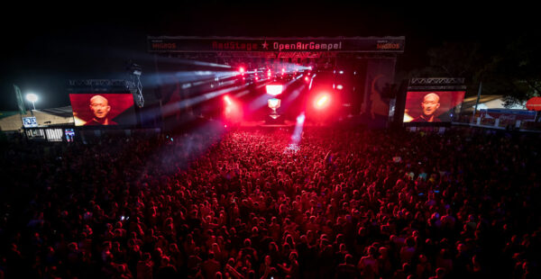 A large crowd of concert-goers at night, immersed in a red-hued stage lighting at an outdoor festival with large screens displaying a performer's face.