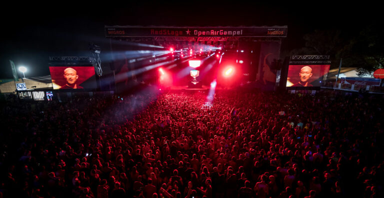 A large crowd of concert-goers at night, immersed in a red-hued stage lighting at an outdoor festival with large screens displaying a performer's face.