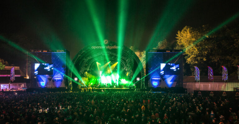 A crowded outdoor music festival at night with a large stage illuminated by green lights and flanked by screens showing visuals, with the words 