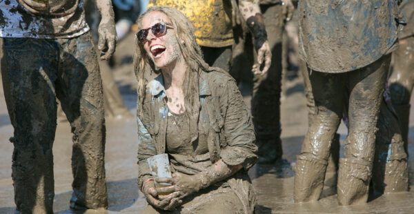 A person smiling and covered in mud, sitting on the ground at a muddy outdoor event, surrounded by other people also coated in mud.