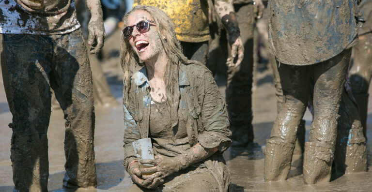 A person smiling and covered in mud, sitting on the ground at a muddy outdoor event, surrounded by other people also coated in mud.