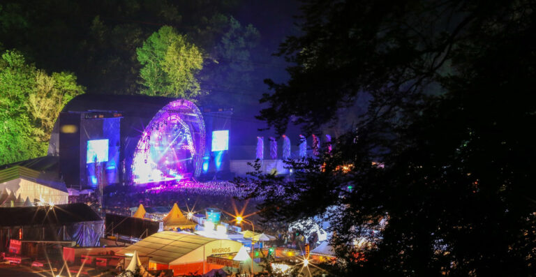 A night view of an outdoor music festival with a large stage illuminated by purple and blue lights, a dense crowd of spectators, and festival tents in the foreground with surrounding trees.