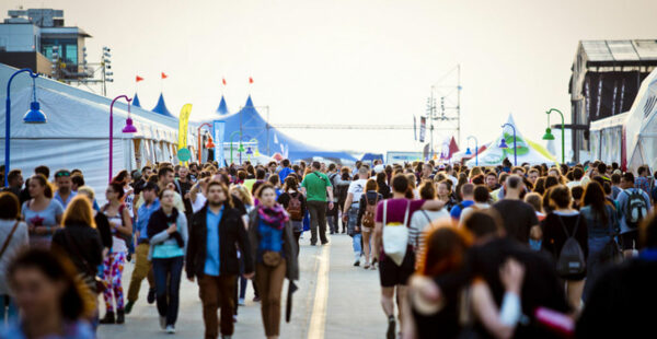 A crowded festival scene with numerous people walking along a wide promenade lined with tents and flags, with a warm sunset light illuminating the scene.