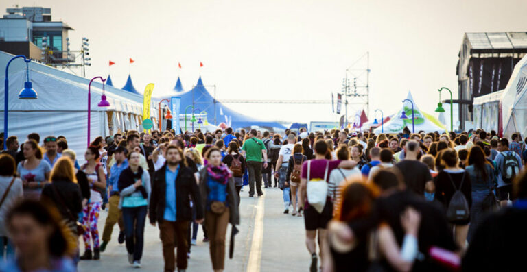 A crowded festival scene with numerous people walking along a wide promenade lined with tents and flags, with a warm sunset light illuminating the scene.