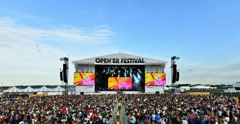 A large crowd of people at the Open'er Festival with a big stage displaying colorful visuals under a clear blue sky.