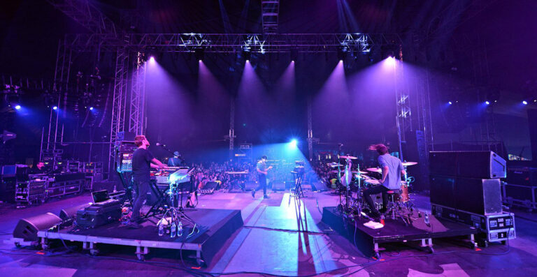 Band performing on stage at a concert with blue stage lighting and audience in the background.