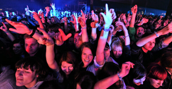Audience at a concert cheering and raising their hands with a colorful stage lighting in the background.