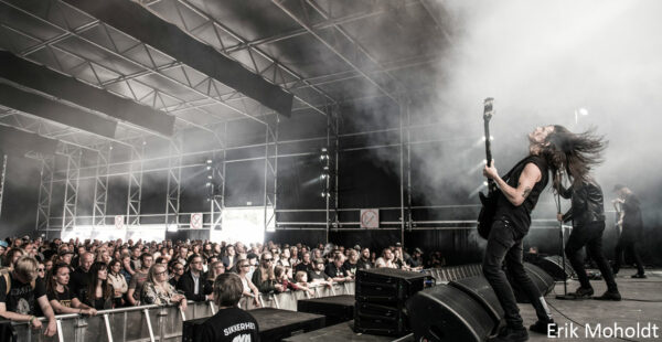 A rock band performs on stage at a music festival, with a guitarist in the foreground playing enthusiastically while hair flies mid-headbang, and an attentive crowd watching amidst stage smoke.