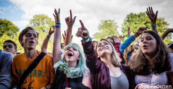 A group of excited young people at an outdoor event, cheering and raising their hands in the air, with trees and sky in the background.