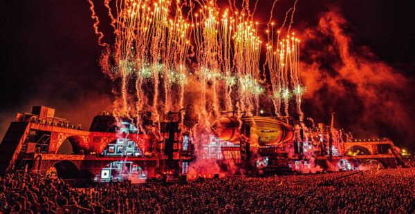 A large outdoor music festival at night with an elaborate stage setup, featuring a vibrant firework display above a massive crowd of spectators.
