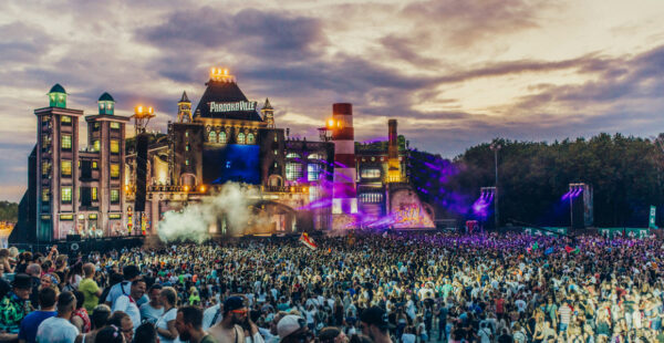 A vast crowd of festival-goers in front of a large, elaborate stage with colorful lights and smoke effects at dusk, with the words 