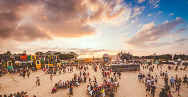 Outdoor music festival scene with a crowd of people, some sitting and others walking, against a backdrop of a dramatic sunset sky. A large 
