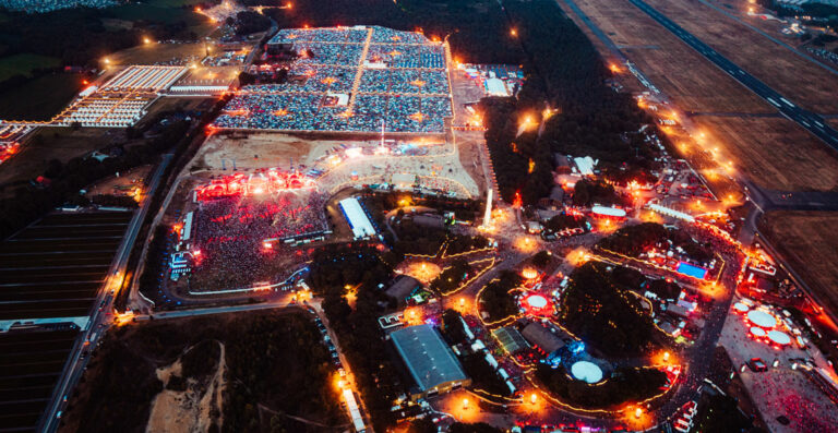 Aerial view of a vibrant outdoor festival at night, showcasing bright lights, multiple stages, and large crowds with adjoining parking areas and access roads.