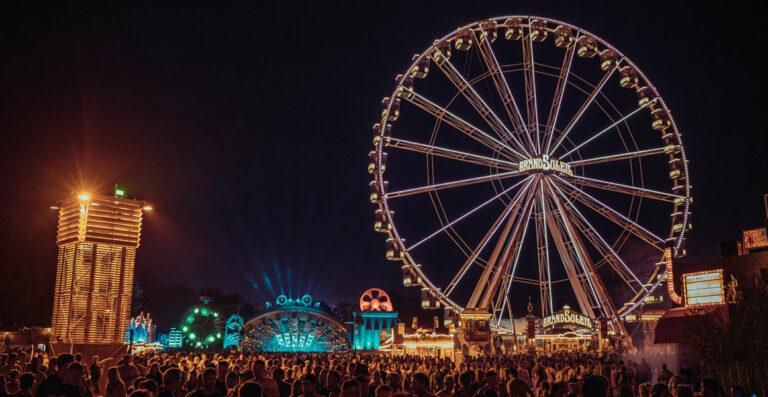 A vibrant night scene at a fair with a large, illuminated Ferris wheel, a crowd of people, and various lighted attractions and booths.