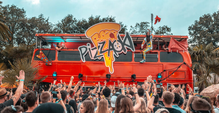A vibrant outdoor music event with a crowd of people cheering and raising their hands towards a red double-decker bus with the word 