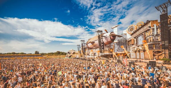 A large crowd of people at an outdoor music festival with a sunny blue sky, standing in front of a stage with elaborate steampunk-themed decoration.