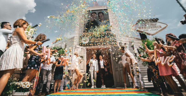 A joyous wedding scene with guests throwing confetti over a newlywed couple exiting a church; the couple is smiling and walking on a rainbow-colored path, while the word 