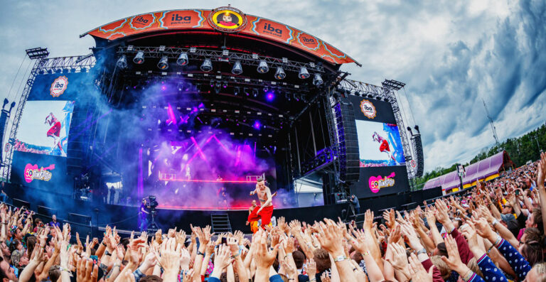 A vibrant outdoor music festival scene with a large crowd of fans raising their hands towards a stage where an artist is performing, with stage screens showing the performer, amid a setting with dramatic purple lighting and a partly cloudy sky.
